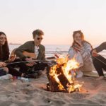 Five young people sitting around a campfire at the beach during sunset playing acoustic guitar and eating sausages
