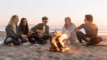 Five young people sitting around a campfire at the beach during sunset playing acoustic guitar and eating sausages