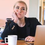 Sad blonde woman dressed in black is sitting in front of a silver macbook air