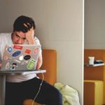 Stressed male student in a white T-shirt sitting in front of a laptop in study room