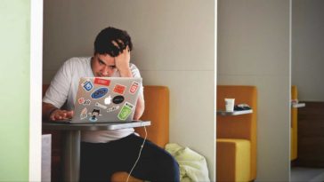 Stressed male student in a white T-shirt sitting in front of a laptop in study room