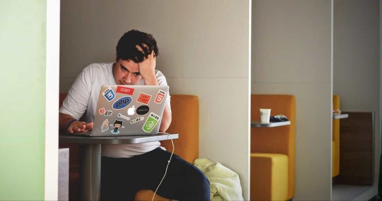 Stressed male student in a white T-shirt sitting in front of a laptop in study room